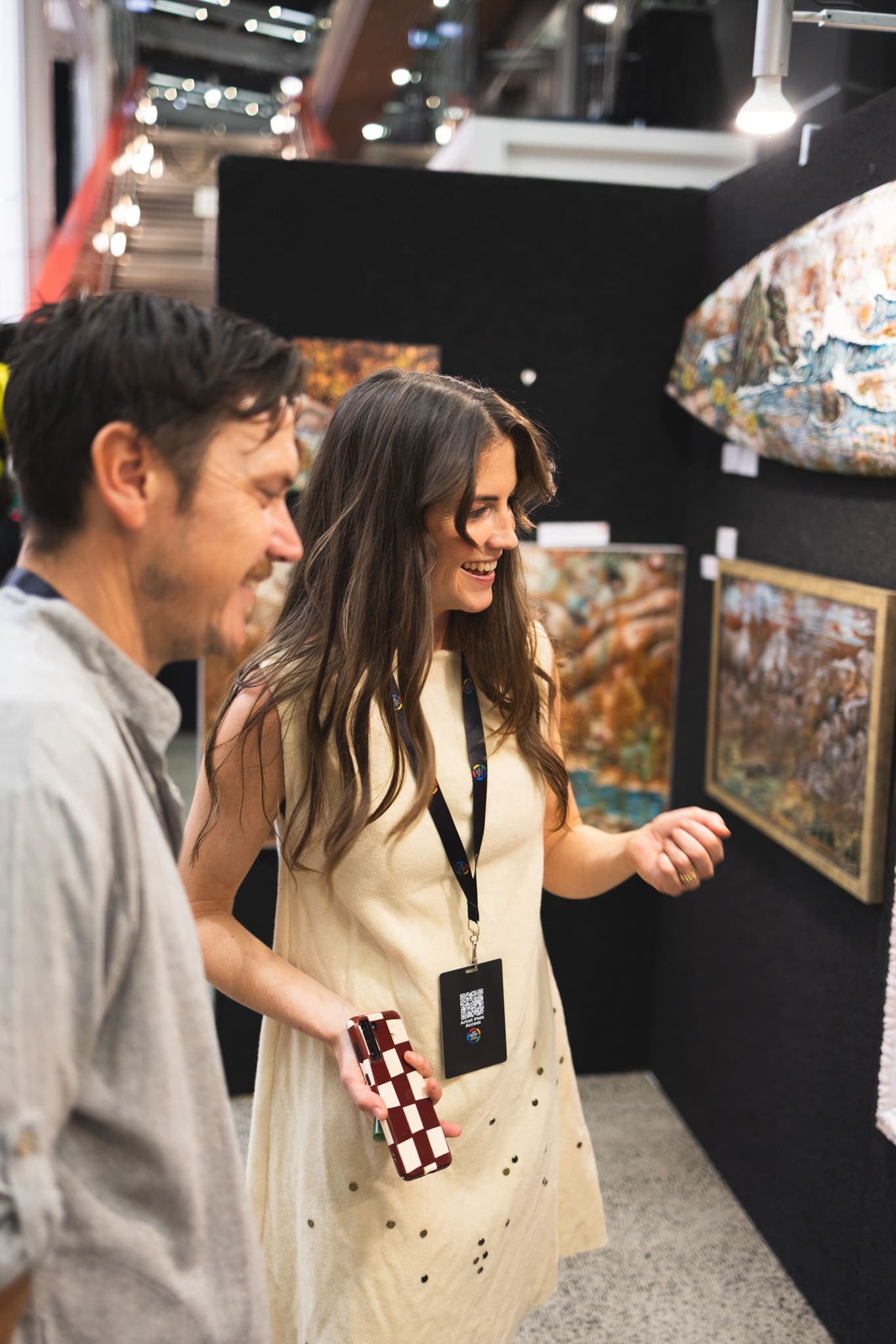 Caitlin laughing with a visitor in her booth, gesturing towards the framed works on the wall