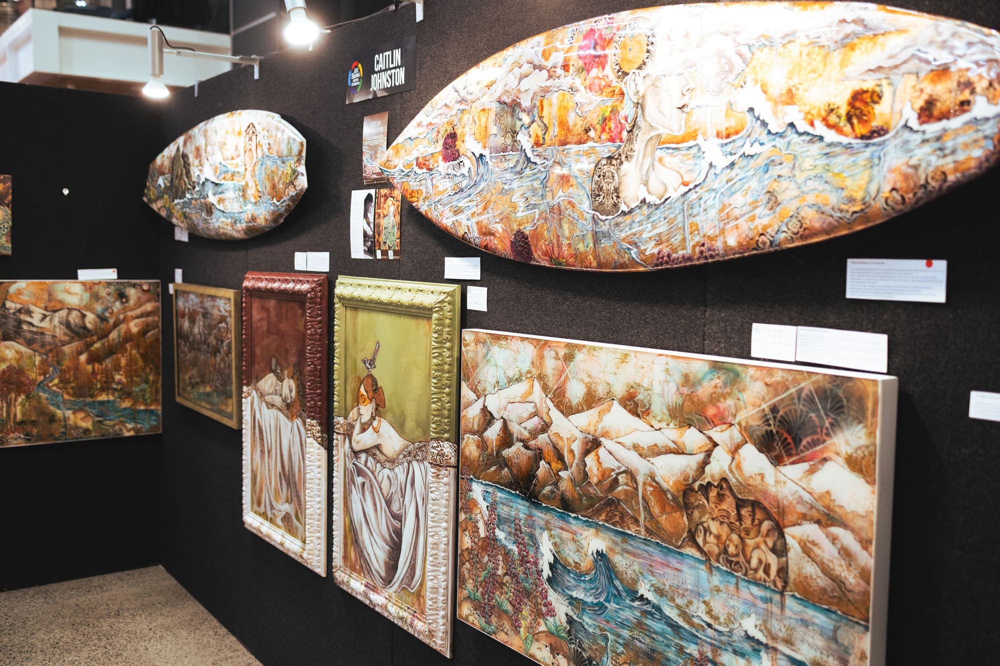 The booth wall seen from the side — a painted surfboard and oval board above framed figure works and a wide painted mountain-and-river panel
